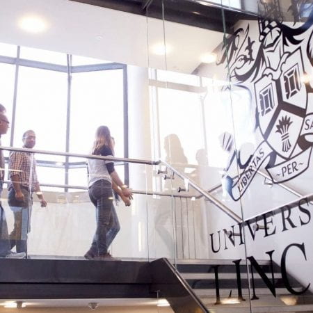 People going downstairs on an University building. There is a glass wall with the University of Lincoln logo on it