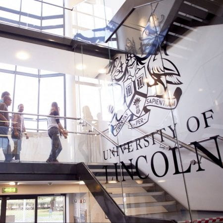 An image of people walking downstairs. The University of Lincoln Logo on a glass wall