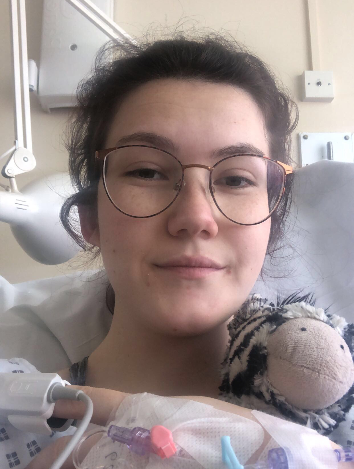 An image of Jo, a white female presenting person with brown hair which has been tied back. Jo has just come out of surgery and is in a hospital gown. In the photo, the cannula in Jo's hand is visible as well as an oximeter on her finger. Jo is also holding a cuddly zebra toy.