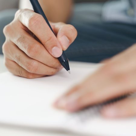 Close up of young man in casual cloth hands writing down on the notepad, notebook using ballpoint pen on the table.