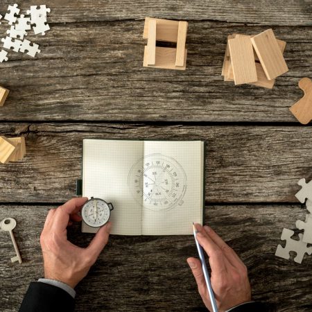 Businessman making plan and business strategy decisions as he sketches a compass he is holding into his notebook. Various cubes, pegs, puzzles and a key lying on his wooden office desk, top view.